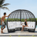 A large black oval wicker lounge chair with white cushions sits on a wooden deck beside a pool, with a man standing on the left and a woman sitting on the right.