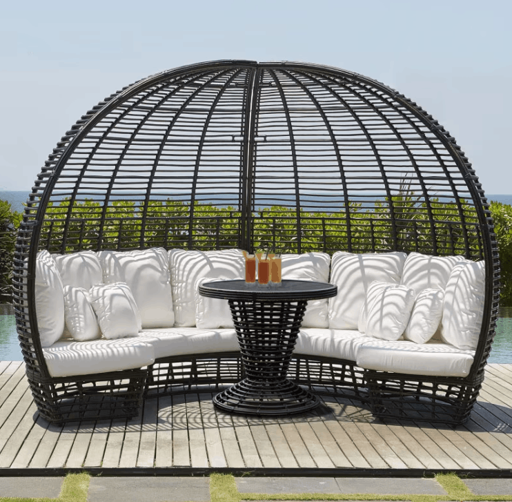 A large oval black wicker daybed with white cushions and a matching round black table, set on a wooden deck with a pool and greenery in the background.