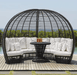 A large oval black wicker daybed with white cushions and a matching round black table, set on a wooden deck with a pool and greenery in the background.