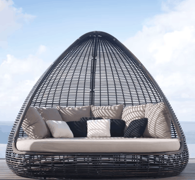 A teardrop-shaped outdoor egg chair with a black woven frame, featuring a thick cream cushion and multiple decorative throw pillows, set against a background of blue sky and ocean.