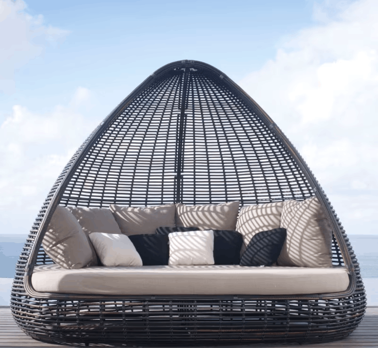 A teardrop-shaped outdoor egg chair with a black woven frame, featuring a thick cream cushion and multiple decorative throw pillows, set against a background of blue sky and ocean.