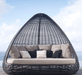 A teardrop-shaped outdoor egg chair with a black woven frame, featuring a thick cream cushion and multiple decorative throw pillows, set against a background of blue sky and ocean.