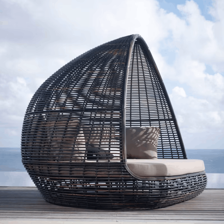 A dark woven egg-shaped outdoor daybed with light beige cushions sits on a wooden deck with the ocean and cloudy sky in the background.