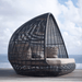A dark woven egg-shaped outdoor daybed with light beige cushions sits on a wooden deck with the ocean and cloudy sky in the background.