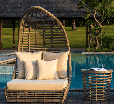 A large woven rattan egg-shaped daybed with cream cushions sits beside a swimming pool on a wooden deck, with a matching woven side table nearby and a resort-style garden background.