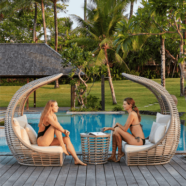 Two women relax in large curved wicker cabana lounge chairs by a tropical swimming pool, with palm trees and a resort-style garden in the background.