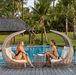 Two women relax in large curved wicker cabana lounge chairs by a tropical swimming pool, with palm trees and a resort-style garden in the background.