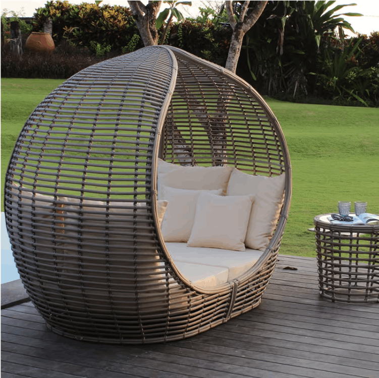 A large teardrop-shaped woven wicker daybed with cream cushions sits on a wooden deck outdoors, with a matching small side table nearby and a green lawn in the background.