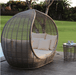 A large teardrop-shaped woven wicker daybed with cream cushions sits on a wooden deck outdoors, with a matching small side table nearby and a green lawn in the background.