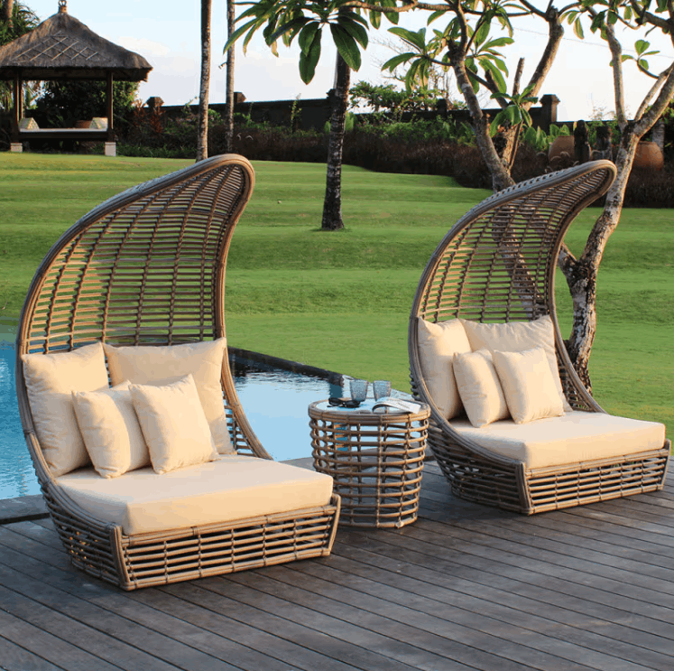 Two high-back woven rattan lounge chairs with cream cushions and a matching round side table on a wooden deck beside a pool, with a tropical garden background.