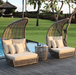 Two high-back woven rattan lounge chairs with cream cushions and a matching round side table on a wooden deck beside a pool, with a tropical garden background.