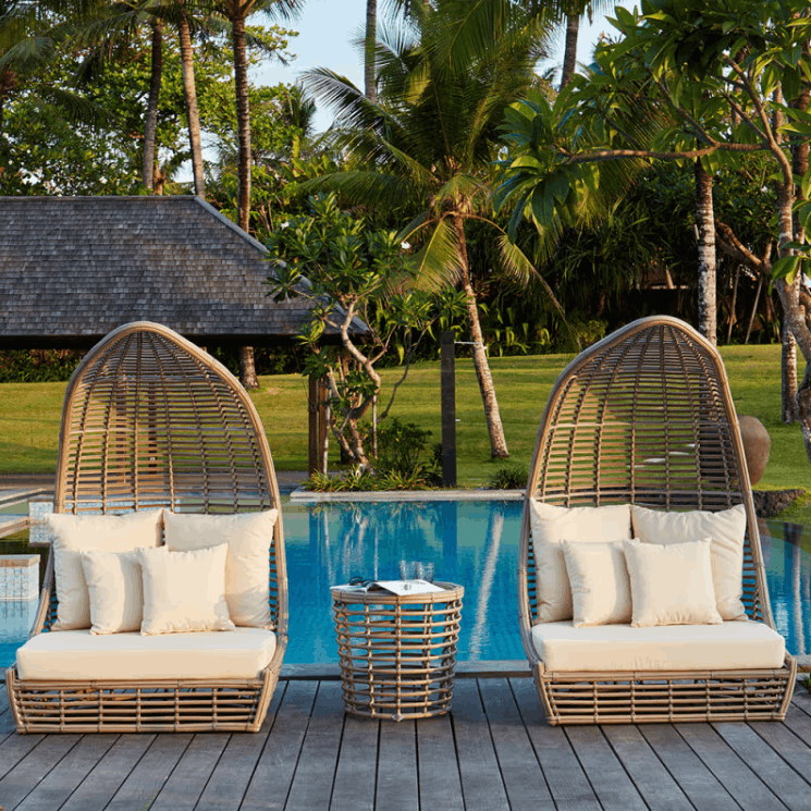 Two tall woven wicker pod lounge chairs with cream cushions are placed on a wooden deck beside a blue swimming pool, with palm trees and a tropical garden in the background.