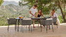 An outdoor dining set with a rectangular dark gray table and four woven gray chairs, shown on a patio with three people dining under trees and mountains in the background.