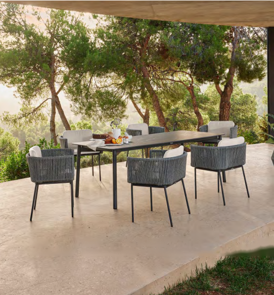 An outdoor dining set consisting of a rectangular dark gray table and six woven gray chairs with white cushions, arranged on a concrete patio with trees in the background.