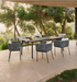 An outdoor dining set consisting of a rectangular dark gray table and six woven gray chairs with white cushions, arranged on a concrete patio with trees in the background.