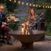 Two people roasting marshmallows over an Arteflame Classic 40 inch corten steel fire pit on a deck at dusk