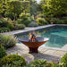 Arteflame Classic 40 inch corten steel fire pit burning poolside on a stone patio surrounded by garden plantings