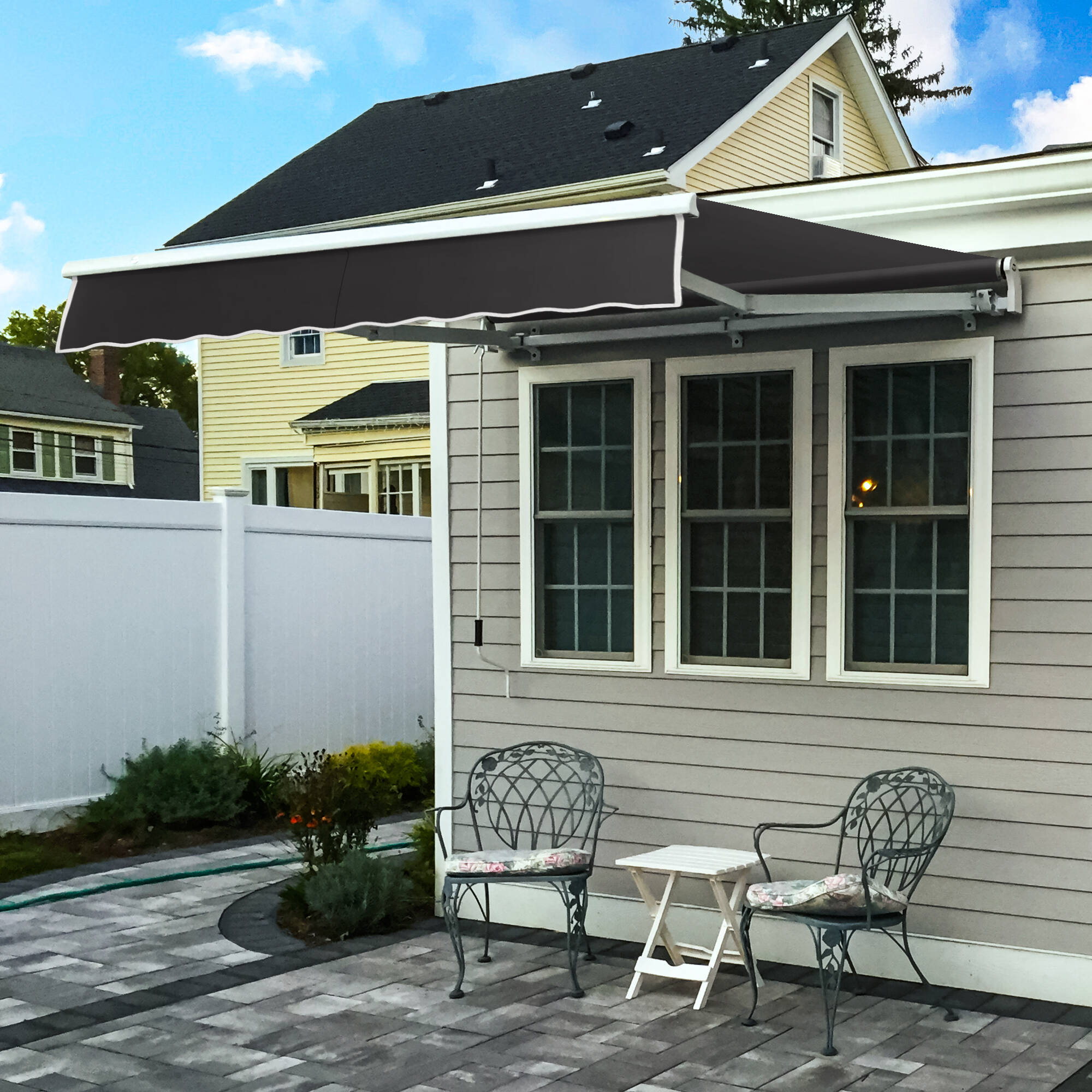 Awntech Maui motorized retractable awning in charcoal acrylic extended over a paver patio with two wrought iron chairs beside a gray clapboard house