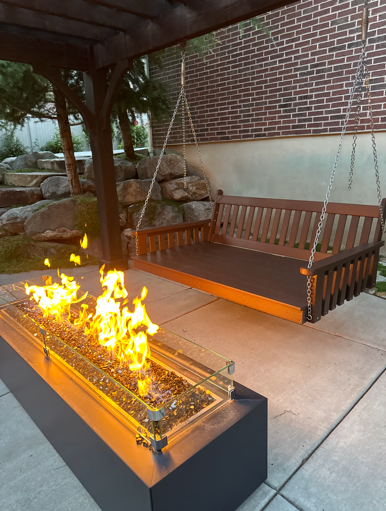 Customer photo: A&L Poly Traditional swing bed in Tudor Brown hung from a backyard pergola, with a lit fire pit in the foreground