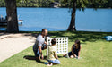 A man and two children playing Four In A Row on a lawn by a lake