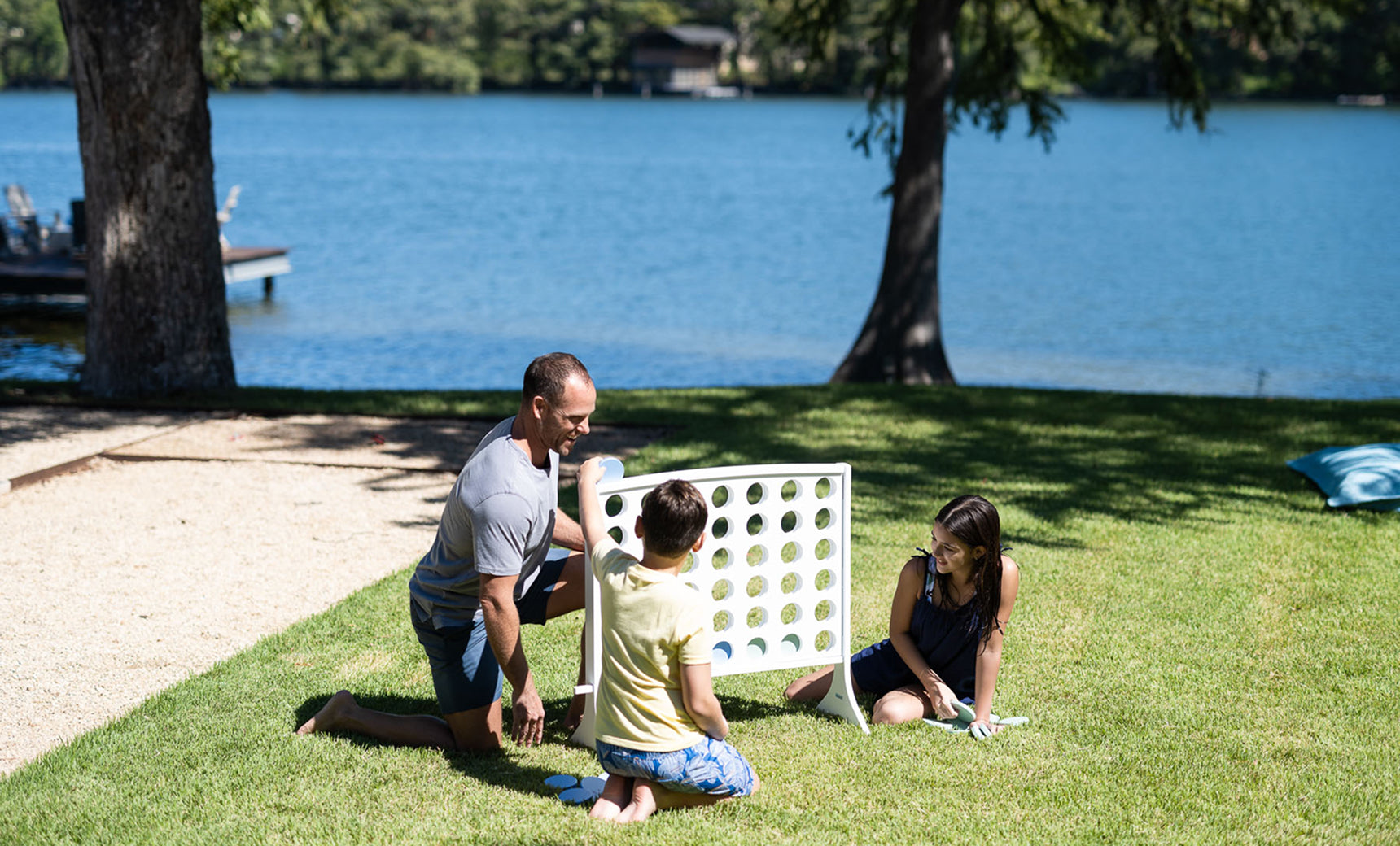 A man and two children playing Four In A Row on a lawn by a lake