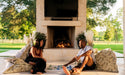 A couple playing giant checkers on floor cushions beside an outdoor fireplace on a sunny patio