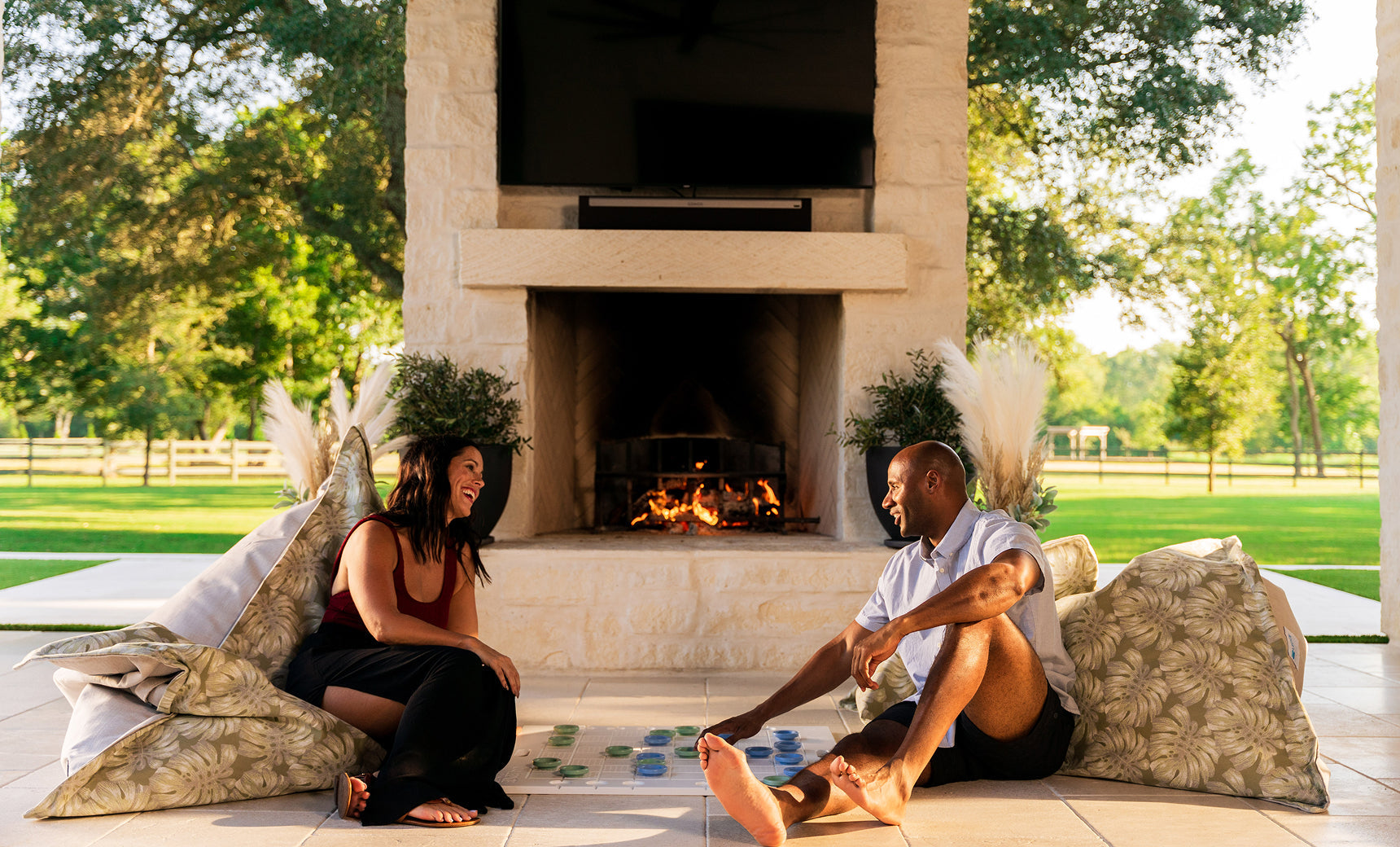 A couple playing giant checkers on floor cushions beside an outdoor fireplace on a sunny patio