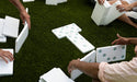Group of people laying out large white dominoes on a green lawn
